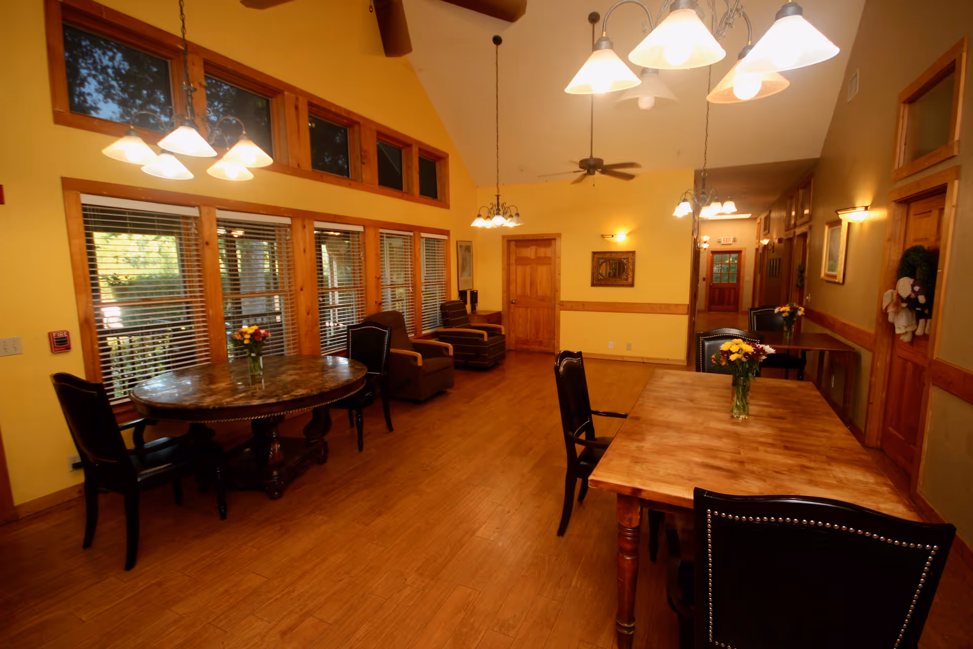 A warm and inviting dining room with wooden floors and yellow walls. The room features two wooden dining tables with chairs, each table decorated with a vase of flowers. Large windows with blinds allow natural light to fill the space. Ceiling fans and multiple light fixtures hang from the ceiling, and there are a few armchairs along the wall.