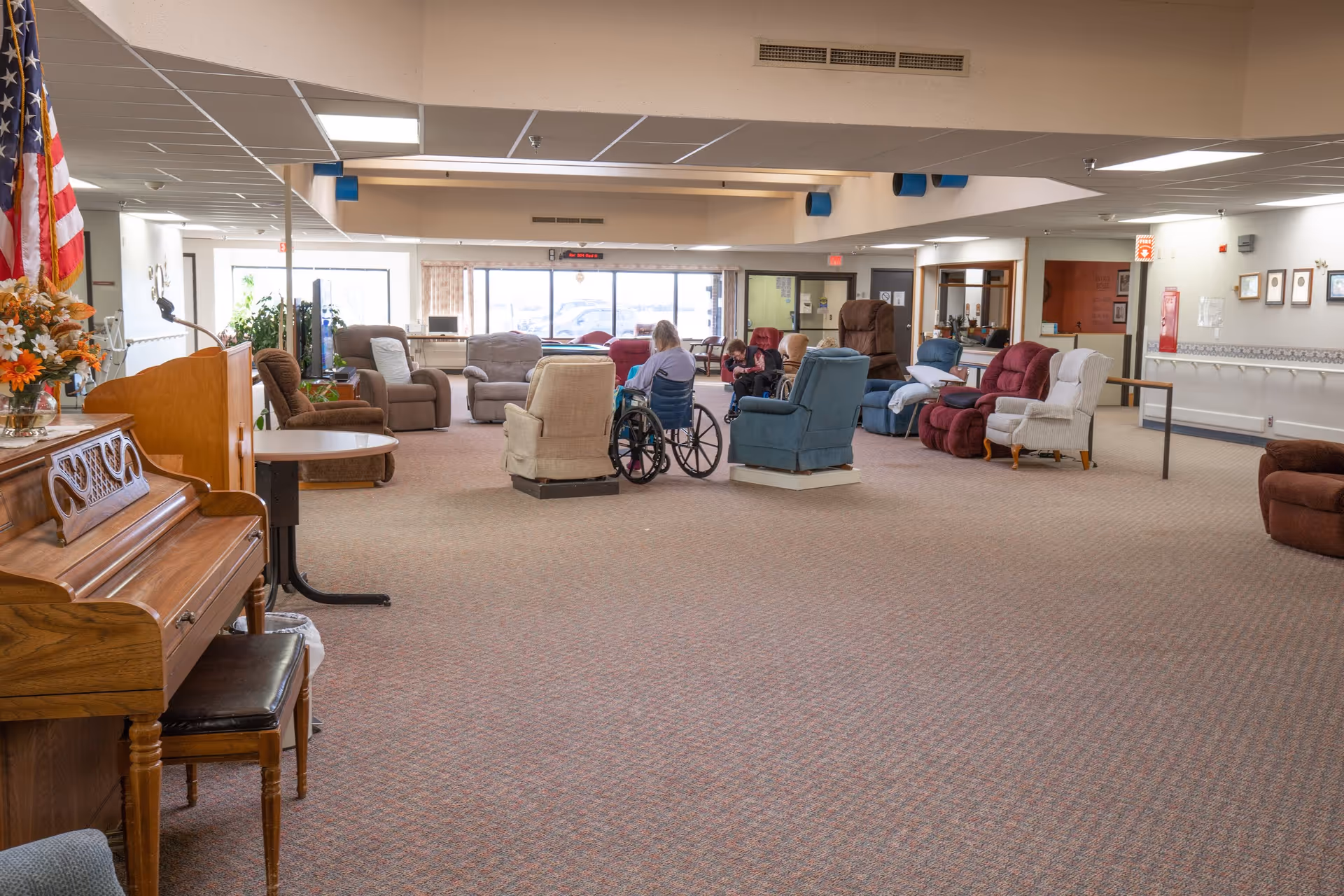 A spacious common area in a senior living facility with various armchairs and recliners arranged in a circle. Two elderly individuals are seated in wheelchairs in the center of the room. The room has a carpeted floor, a piano with a bench on the left, an American flag, and large windows letting in natural light at the far end.