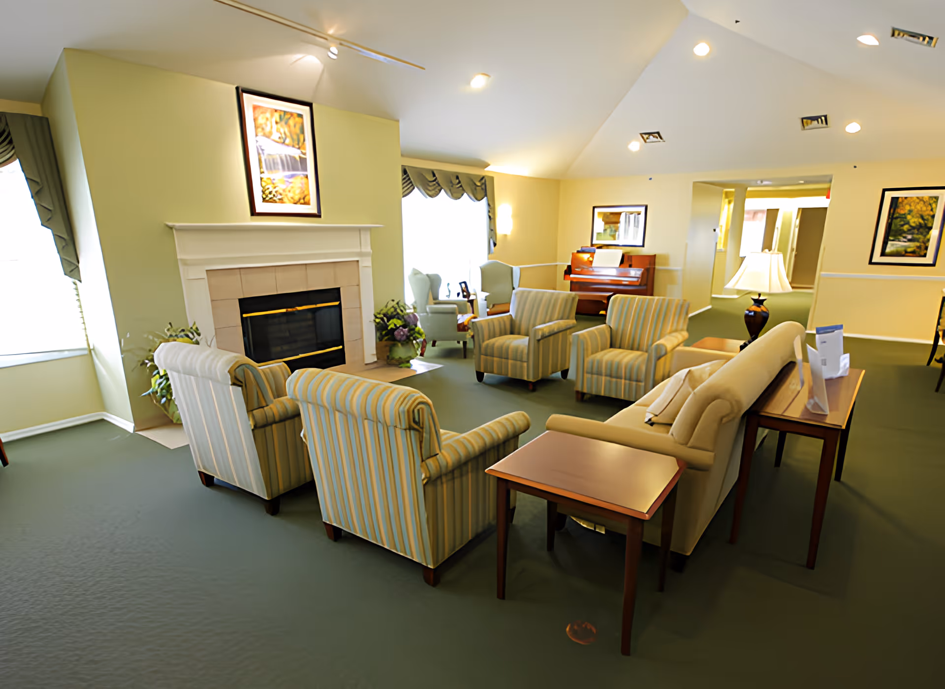 A cozy living room area in a senior living facility with a fireplace, several striped armchairs, a beige sofa, side tables, a lamp, and a piano in the background. The room is well-lit with natural light from windows and ceiling lights, decorated with framed artwork and plants.