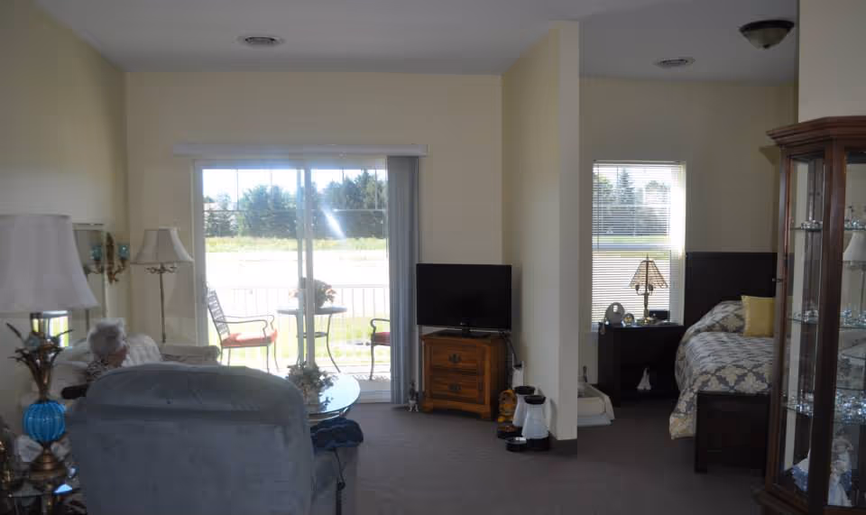 Interior view of a senior living facility room showing a living area with chairs, a small table, and a television on a wooden stand. To the right, there is a bedroom area with a bed, nightstand, and lamp. A glass cabinet with decorative items is visible on the far right. Sliding glass doors lead to an outdoor patio with chairs and a small table.