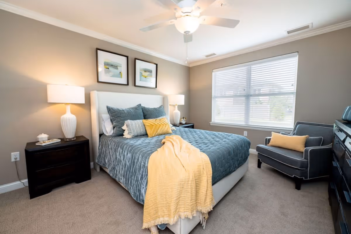 A neatly made bed with a blue quilt and multiple pillows, including a yellow one, in a bedroom with beige walls. There are two framed abstract artworks above the bed, two bedside tables with lamps, a window with blinds, a ceiling fan, a gray armchair with a yellow cushion, and a dark dresser.