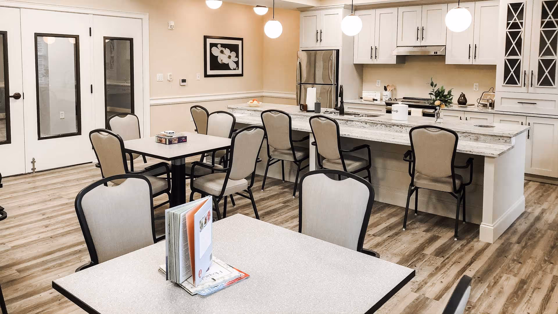 A bright and clean communal dining area with several tables and chairs, adjacent to a modern kitchen featuring white cabinets, a stainless steel refrigerator, and a large island with seating. The room has wood flooring, pendant lights, and a framed floral picture on the wall.