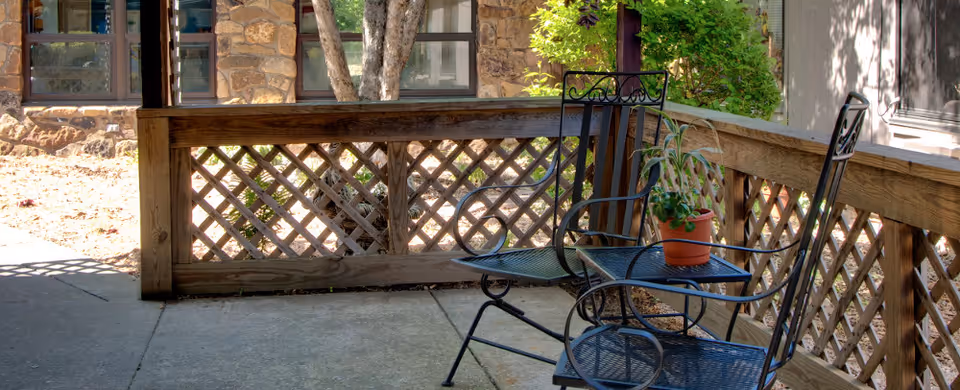 Outdoor patio area with two black metal chairs and a small matching table holding a potted plant. The patio is enclosed by a wooden lattice fence and is adjacent to a stone building with windows and greenery.