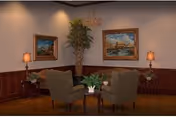 A cozy sitting area with two upholstered armchairs facing each other around a small wooden table with a flower arrangement. The room features wood paneling on the lower half of the walls, two framed paintings, two table lamps on side tables, and a tall potted plant in the corner.