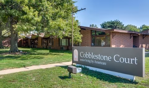 Brick senior living building exterior with a 'Cobblestone Court' sign on the lawn and trees.
