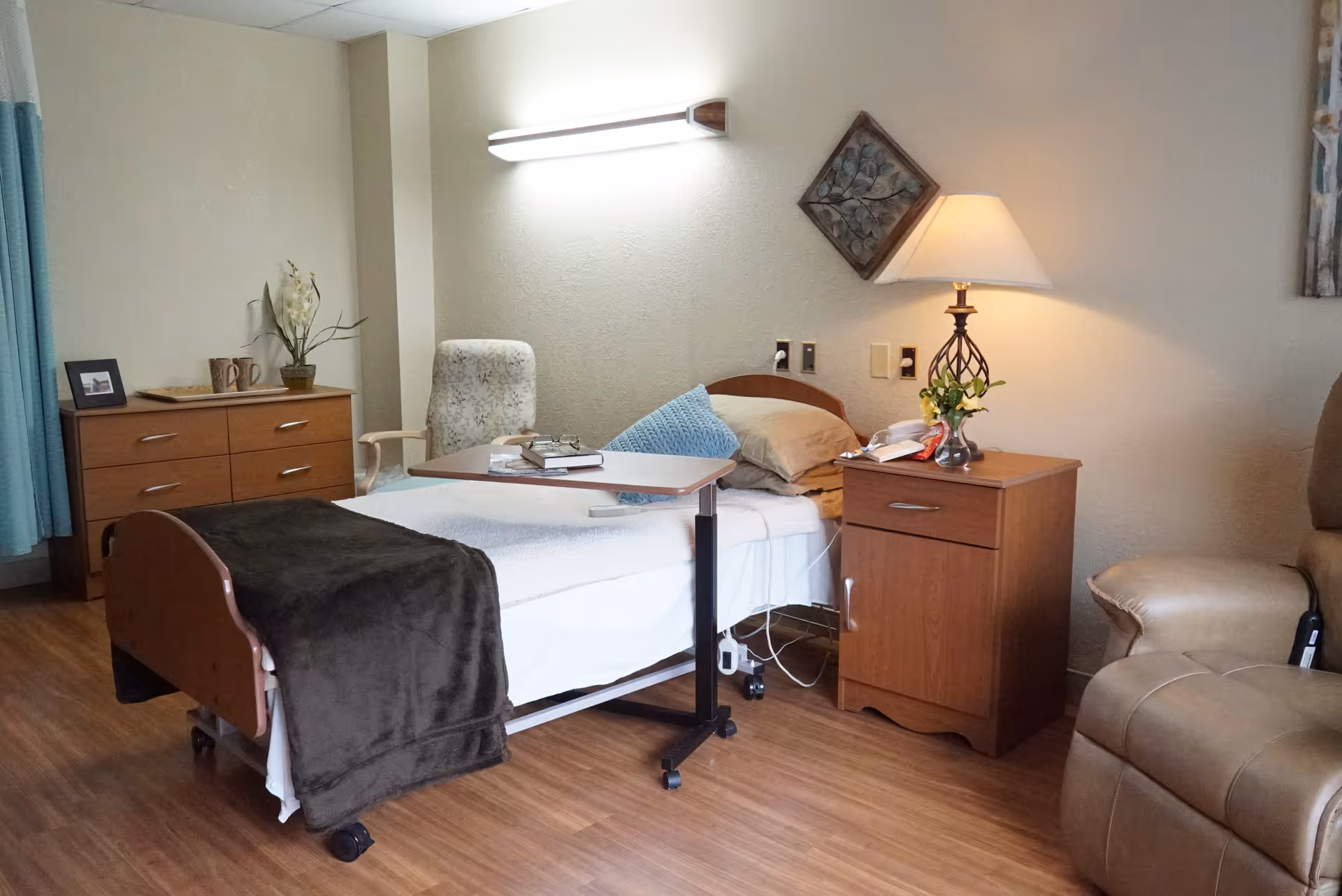 A cozy bedroom in a senior living facility featuring a single bed with a brown blanket and blue pillow, a wooden bedside table with a lamp and small plant, a wooden dresser with decorative items, a comfortable armchair, and a rolling overbed table with books on it. The room has wooden flooring and soft lighting.
