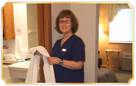 A woman wearing navy blue scrubs and glasses is holding a white towel in a room that appears to be part of a senior living facility. Behind her is a sink with a mirror and a towel ring, and in the background, there is a bed, a side table with a lamp, a window with blinds, and an air conditioning unit.
