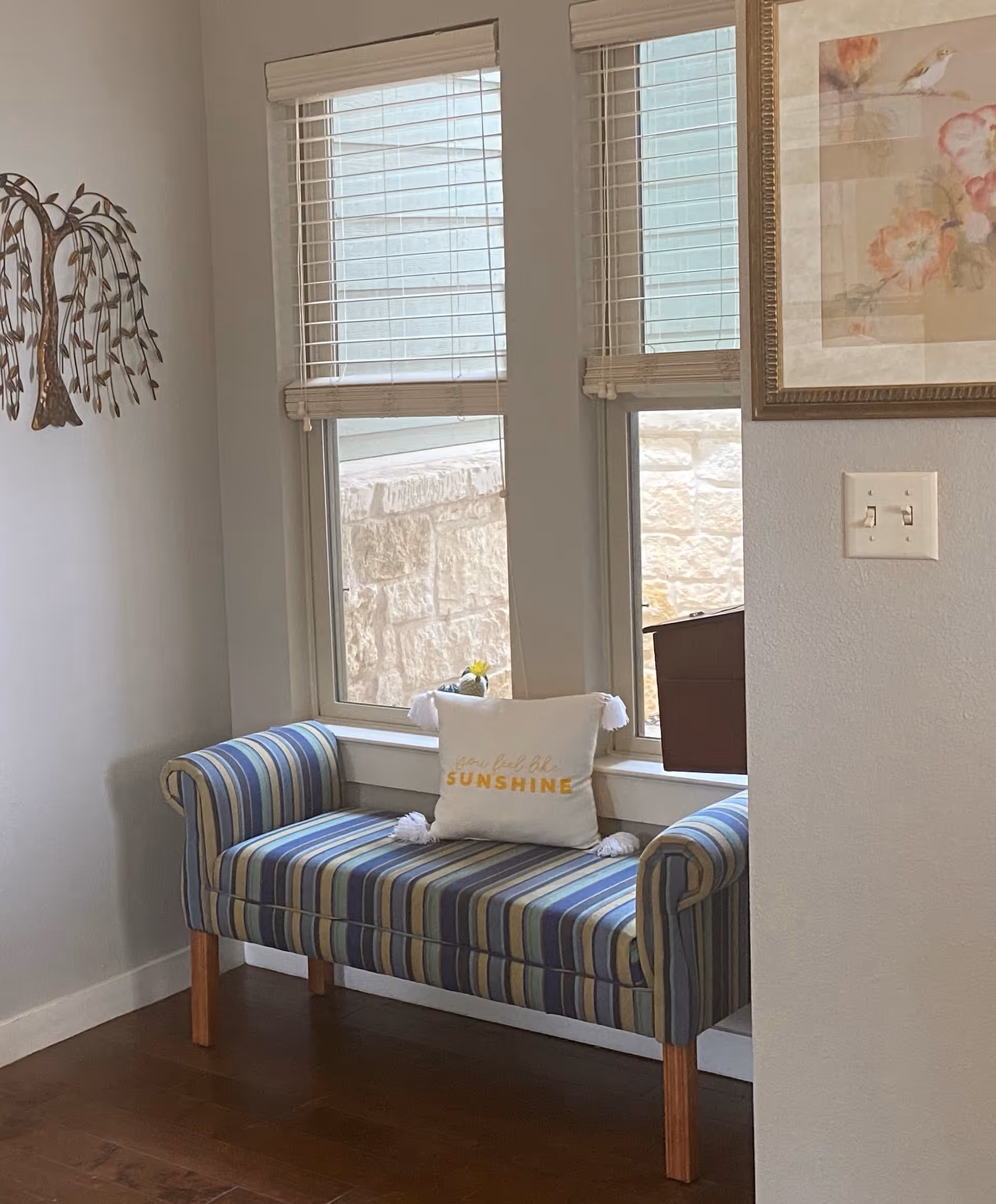 A cozy corner with a striped upholstered bench featuring blue, green, and beige tones, placed against a wall with two windows. A white decorative pillow with the words 'you feel like SUNSHINE' in yellow is on the bench. The wall has a metal tree wall art on the left and a framed floral painting on the right. There is a light switch on the wall near the painting.