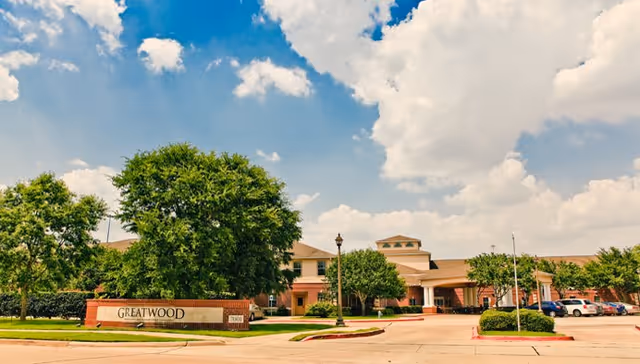 Front exterior of the Greatwood senior living building with a driveway, landscaped trees, and a large entrance sign.