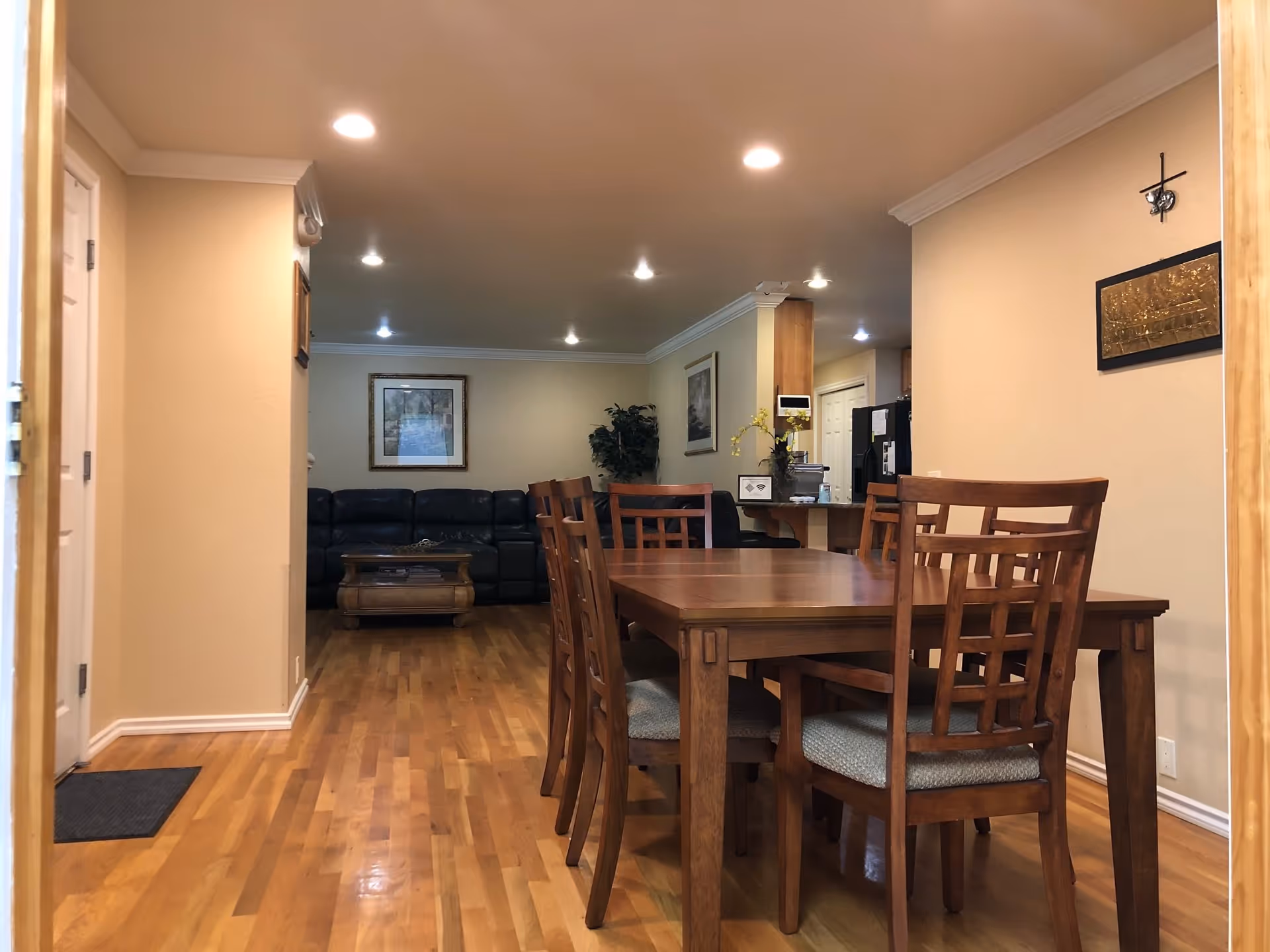 Wooden dining table and chairs in an open-plan room with hardwood floors, recessed lighting, and a living room sofa visible in the background.