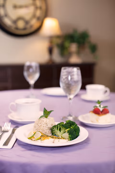 A plated meal of rice, broccoli and fish on a lavender tablecloth with water glasses, teacups and a blurred dining room background.