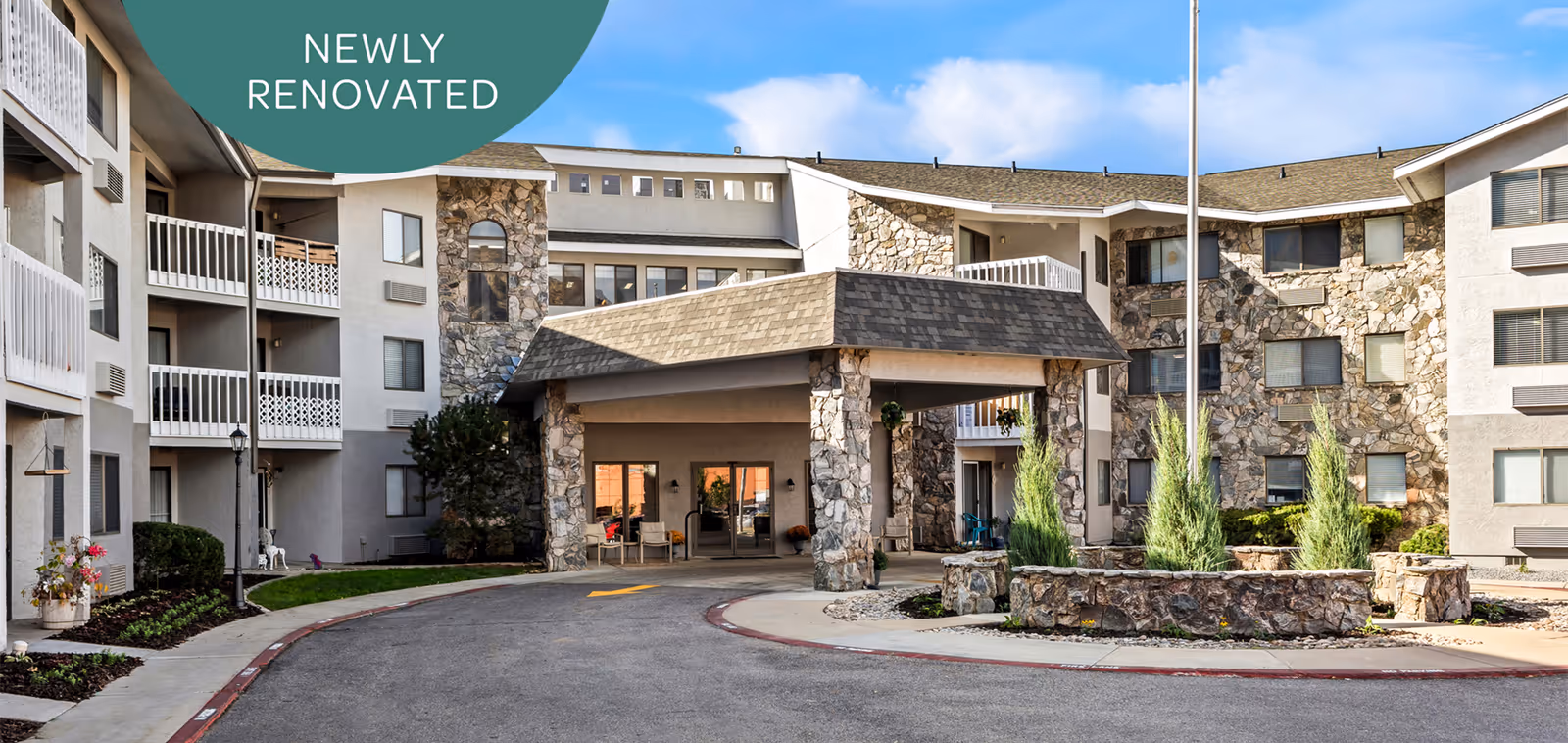 Exterior view of a multi-story retirement community building with stone and beige facade, featuring balconies and a covered entrance driveway. The sky is clear and blue, and there is landscaping with shrubs and flowers around the building. A green circular graphic in the top left corner says 'NEWLY RENOVATED'.