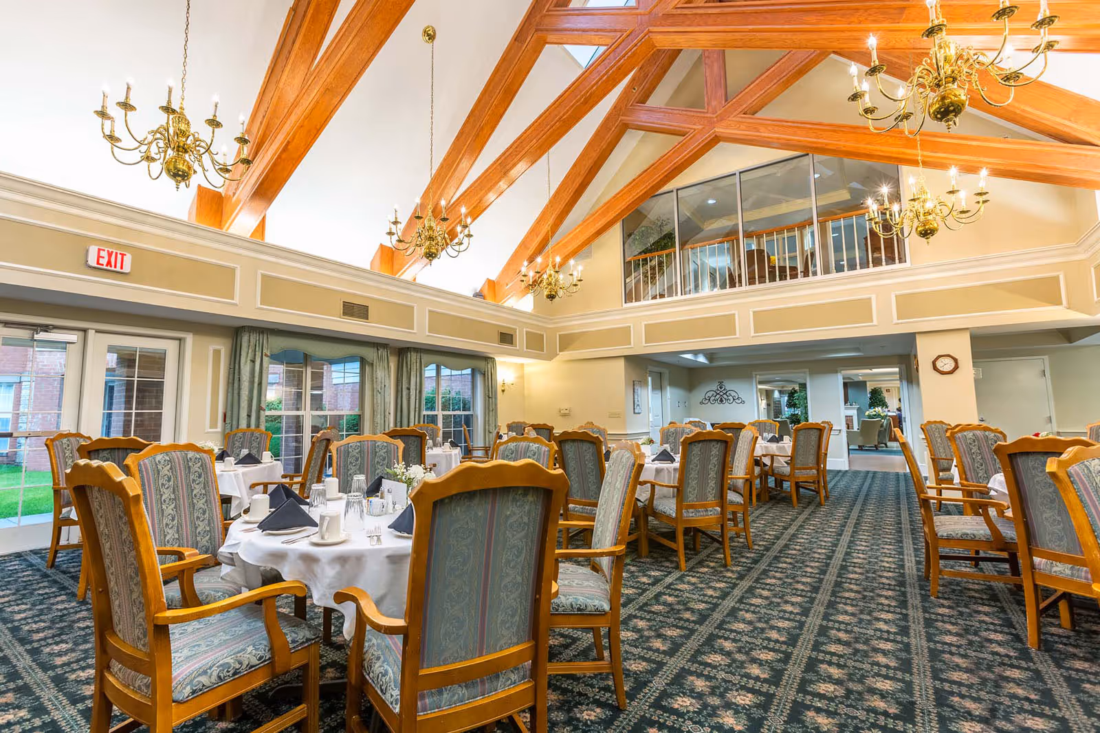 Spacious dining room with tables set for service, upholstered wooden chairs, chandeliers, and exposed wooden ceiling beams.