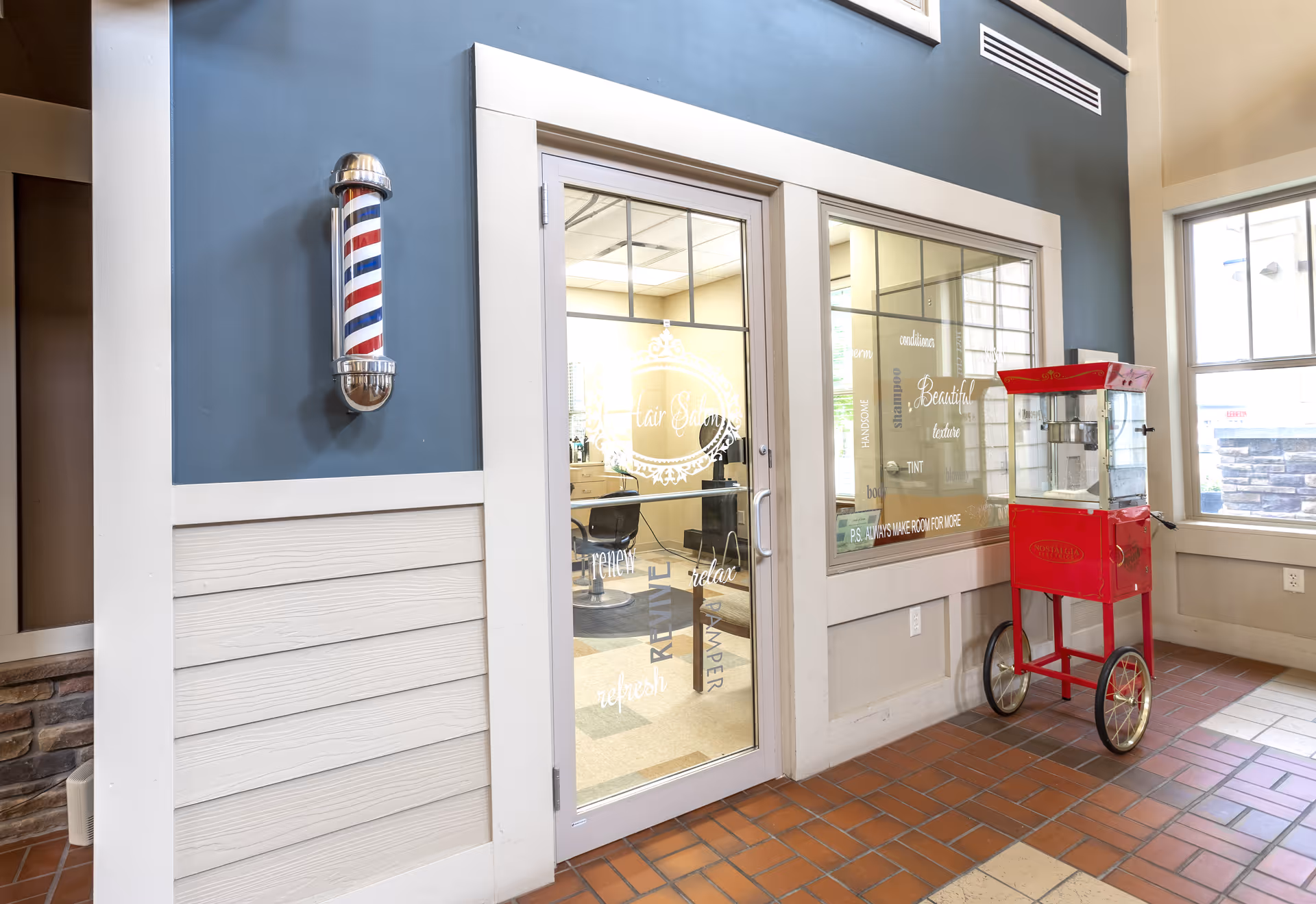 Interior view of a senior living facility showing the entrance to a hair salon with glass doors and windows. A traditional barber pole is mounted on the blue wall to the left of the door. To the right, there is a red vintage-style popcorn machine on wheels. The floor is tiled with a mix of brown and beige tiles, and large windows allow natural light to fill the space.