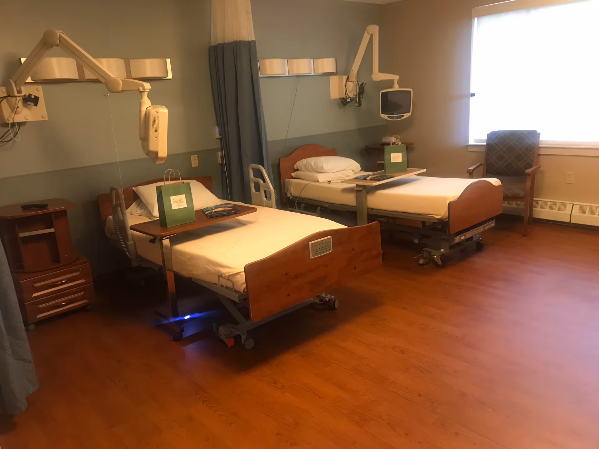 A shared patient room with two hospital beds, over-bed tables, mounted medical equipment, and a chair by a window.