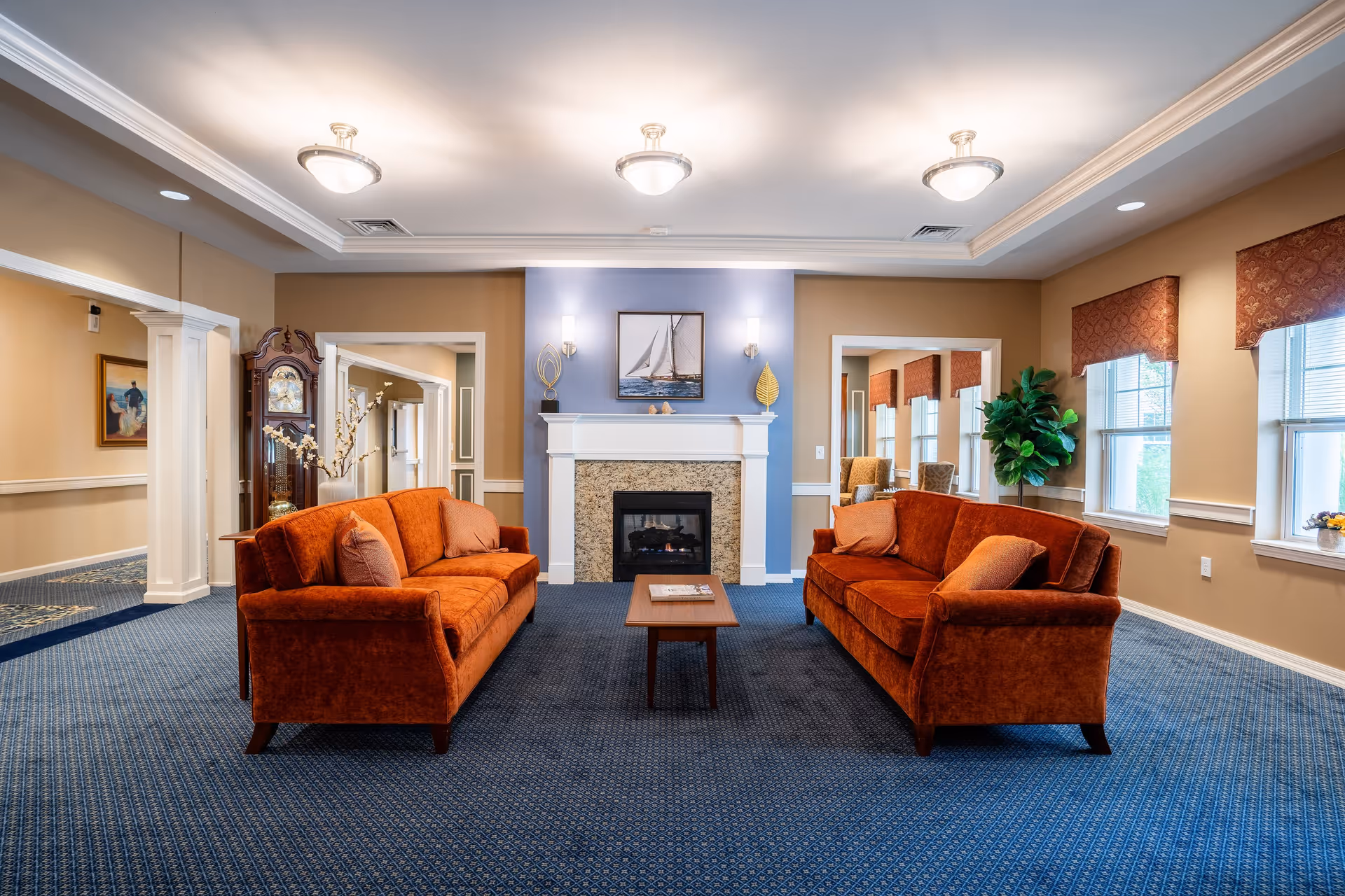A cozy living room with two orange sofas facing each other across a wooden coffee table, a fireplace with a sailboat painting above it, and large windows with patterned valances letting in natural light. The room has blue carpet and beige walls with white trim.
