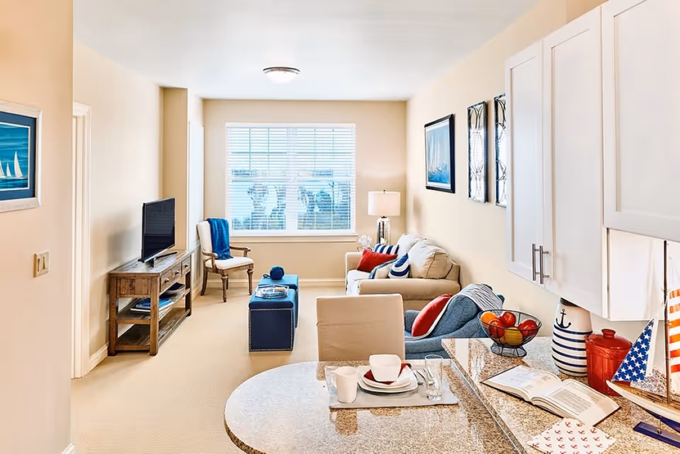 A bright and cozy living room with beige walls and carpet, featuring a beige sofa with colorful pillows, a blue armchair, a wooden TV stand with a flat-screen TV, a window with blinds, and a small dining area with a round table set with dishes and cups. Decorative items include framed nautical-themed artwork, a bowl of fruit, and a model sailboat on the kitchen counter.
