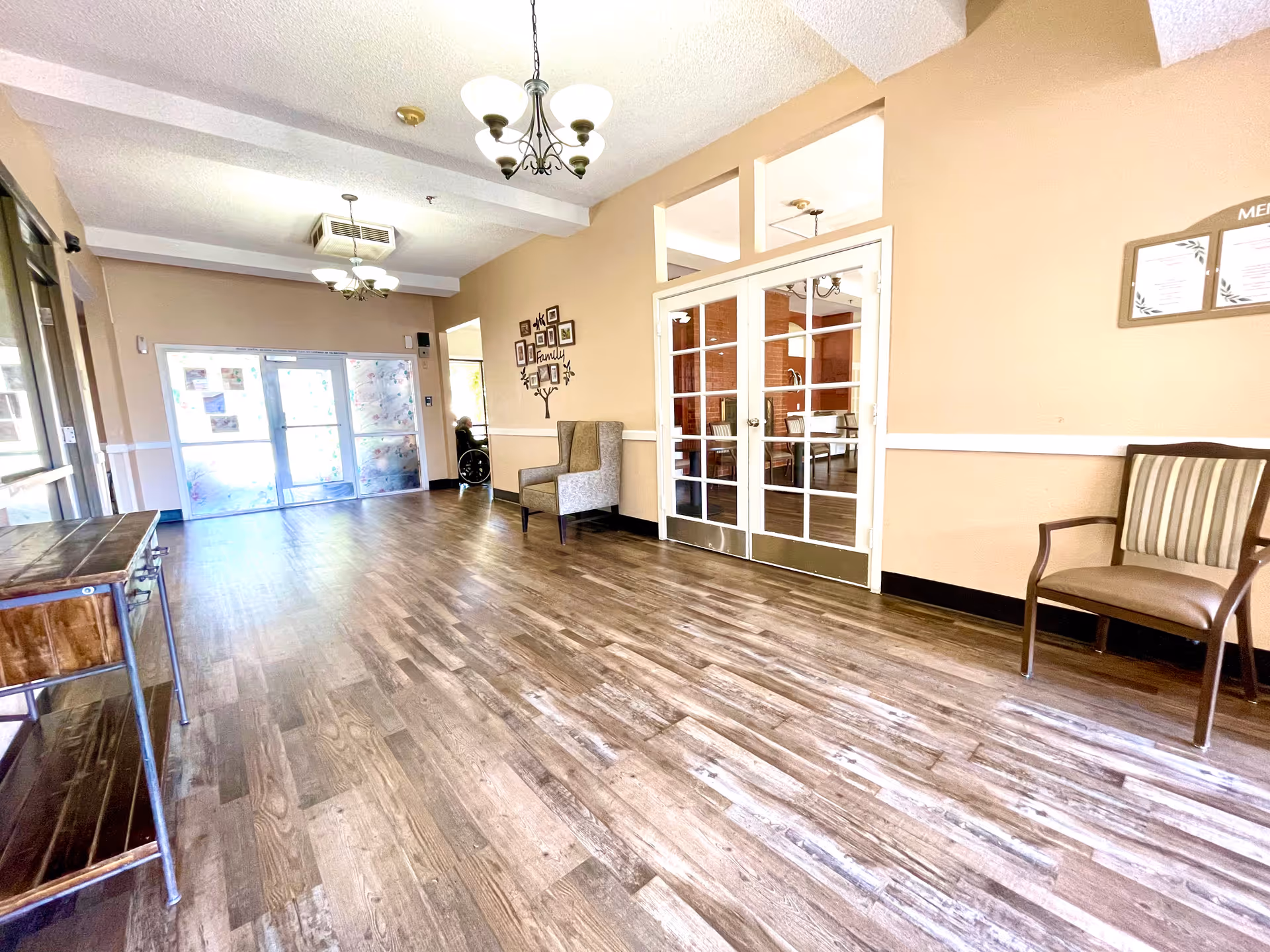 A spacious hallway in a senior living facility with wooden flooring, beige walls, and white trim. There are two chandeliers hanging from the ceiling, a wooden console table on the left, and two chairs on the right side. Double glass doors lead to another room with tables and chairs. A person in a wheelchair is visible near the entrance doors at the far end of the hallway.