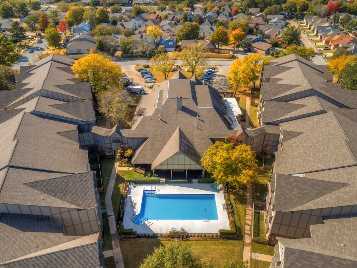 Aerial view of Country Club At Woodland Hills senior living facility showing multiple connected buildings surrounding a central outdoor swimming pool. The area is landscaped with trees displaying autumn colors and a parking lot is visible in the background along with nearby residential houses.