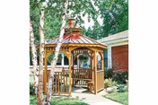 A wooden gazebo with a copper-colored roof situated in a garden area with trees and greenery, adjacent to a brick building.