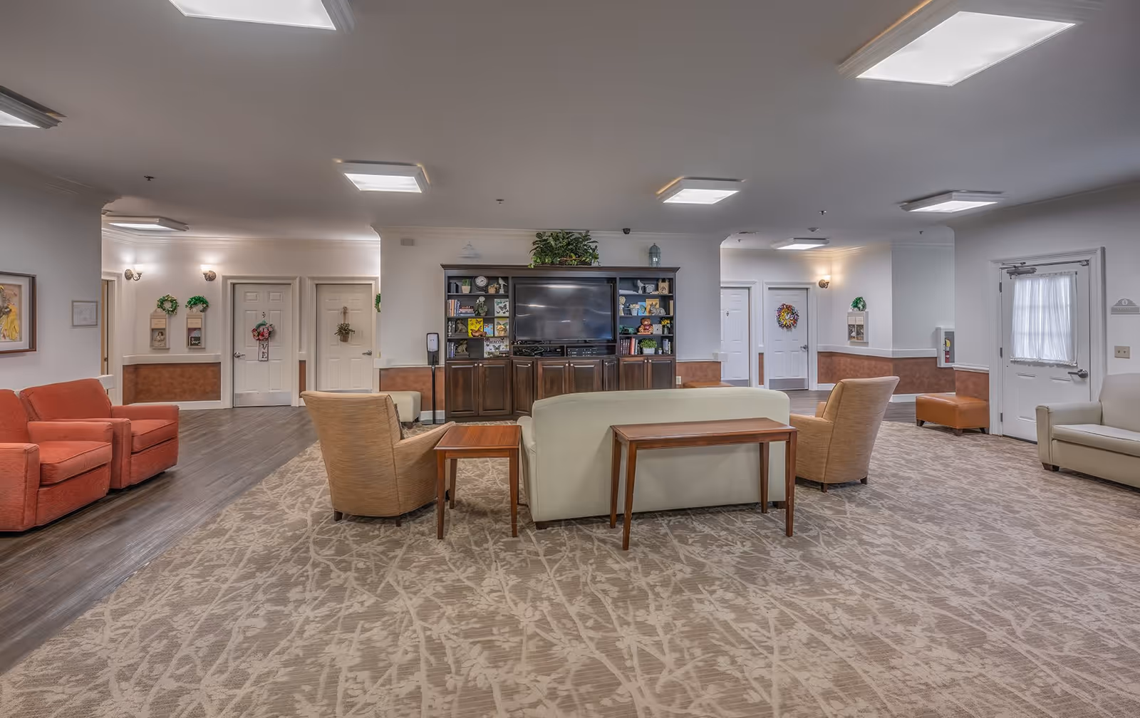 A spacious and well-lit common living area in a senior living facility featuring a large TV on a wooden entertainment center with shelves displaying decorative items. The room has a beige patterned carpet, several comfortable chairs and sofas arranged around the TV, and doors leading to other rooms. The walls are decorated with wreaths and framed pictures, and there is a hand sanitizer dispenser near the TV unit.