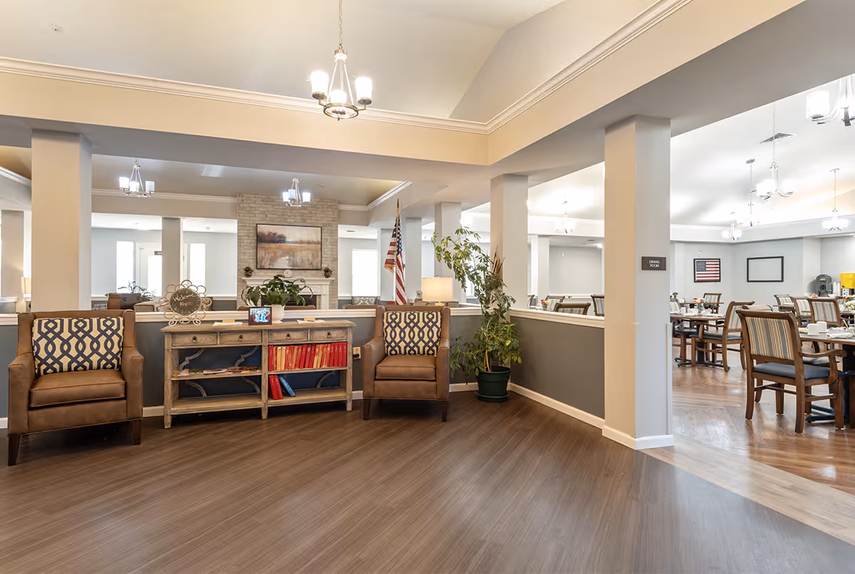 A spacious and well-lit senior living facility common area with two brown armchairs featuring patterned cushions, a wooden console table with books and decorative items, a potted plant, and an American flag. In the background, there is a dining area with multiple tables and chairs, a fireplace with a landscape painting above it, and several ceiling lights.