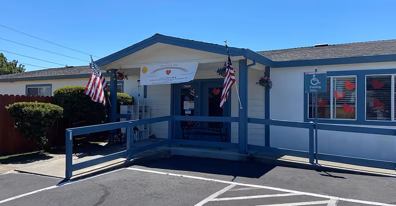 Exterior front view of The Greenhills Care Home building with a wheelchair accessible ramp, two American flags, a banner above the entrance, and a handicapped parking sign. The windows have red heart decorations.