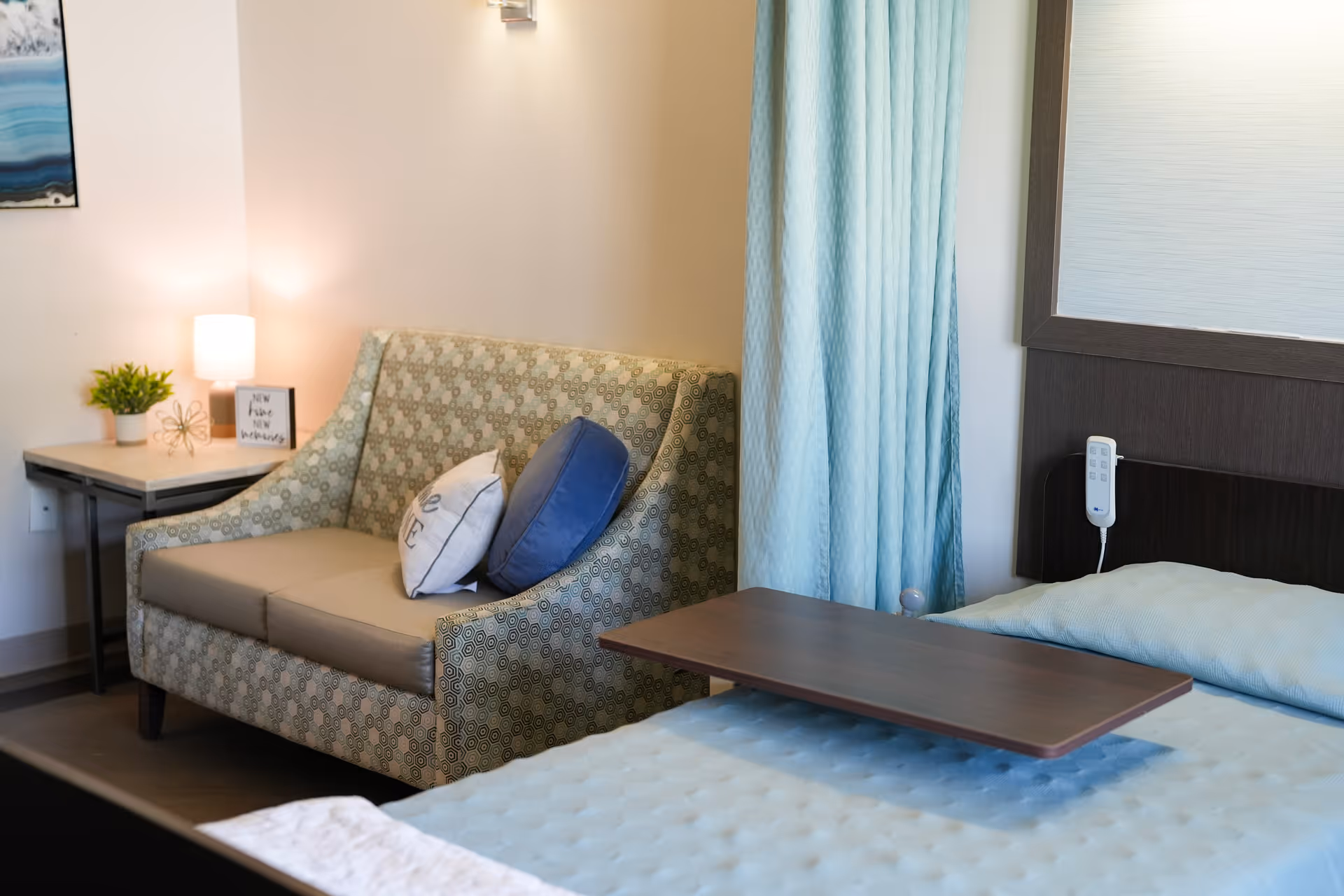 A cozy room featuring a patterned loveseat with two decorative pillows, a small side table with a lamp, a plant, and a framed sign. Next to the loveseat is a bed with a light blue quilted blanket and an adjustable overbed table. Light blue curtains hang behind the bed.