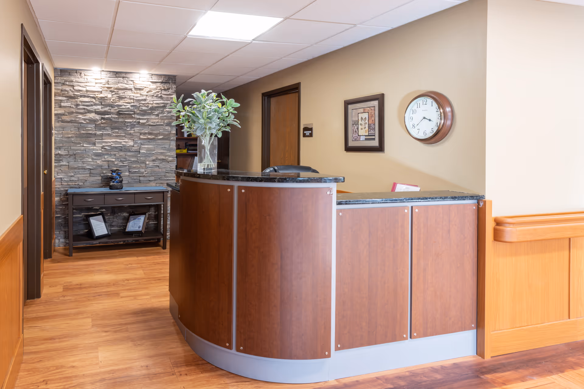 Reception area with a curved wooden desk topped with a black countertop, a vase with green leafy plants, a wall clock, framed artwork, and a stone accent wall with a small table holding decorative items and framed certificates.