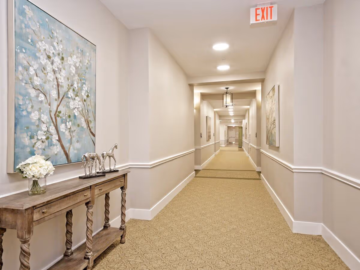 A well-lit hallway in a senior living facility with beige walls and carpeted floor. The hallway features a wooden console table on the left side adorned with a vase of white flowers and two decorative horse figurines. Large floral paintings hang on the walls, and ceiling lights illuminate the corridor. An exit sign is visible on the ceiling near the foreground.