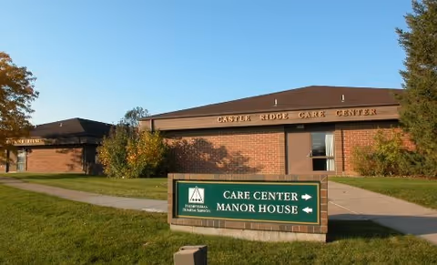 Exterior view of Castle Ridge Care Center, a single-story brick building with a brown roof. In front of the building is a green sign with white text and arrows indicating directions to the Care Center and Manor House. The sky is clear and blue, and there is green grass and some trees around the building.