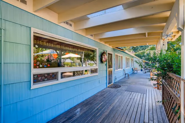 Covered outdoor walkway alongside a light blue building with large windows and a door decorated with a wreath. There are benches along the wall and plants on the right side, with trees and umbrellas visible in the background.
