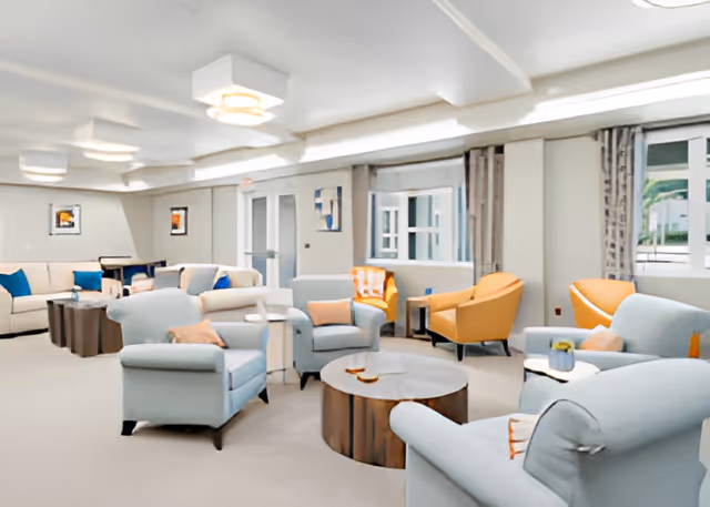 A spacious and well-lit senior living common area featuring multiple comfortable armchairs and sofas in light blue and orange tones arranged around circular wooden coffee tables. The room has large windows with curtains, white walls, and ceiling lights, creating a welcoming and relaxing atmosphere.