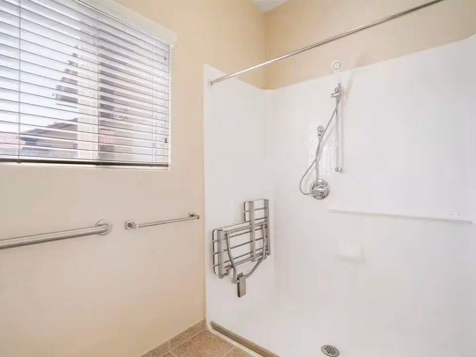 Accessible shower stall with grab bars, handheld showerhead, fold-down seat and a window with blinds.