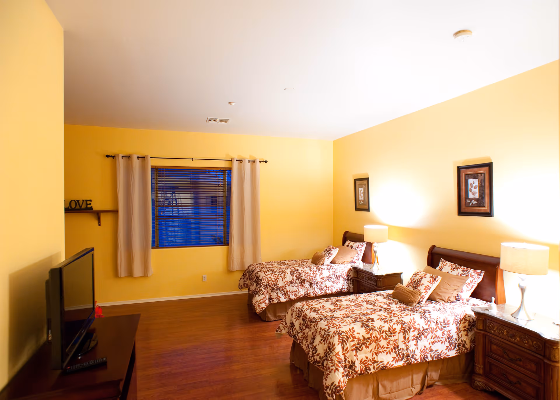 A warmly lit bedroom with two twin beds featuring matching floral bedspreads. Each bed has pillows and a nightstand with a lamp. The walls are painted yellow, and there are two framed pictures hanging above the beds. A window with beige curtains and blinds is on the far wall. A television sits on a wooden stand to the left.