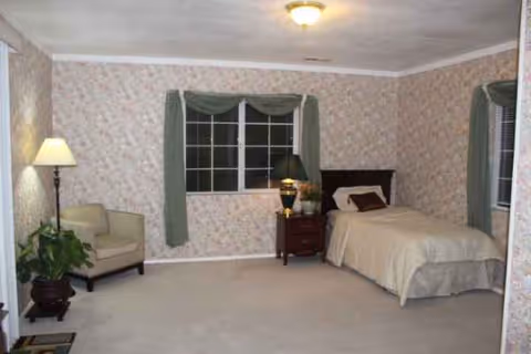 A bedroom with a bed, nightstand, lamp, chair, and floral wallpaper.