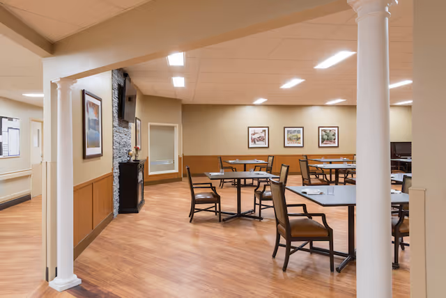 A spacious dining area in a senior living facility with wooden floors, several tables and chairs arranged neatly, beige walls with framed artwork, a stone fireplace with a mounted TV above it, and white columns framing the entrance.