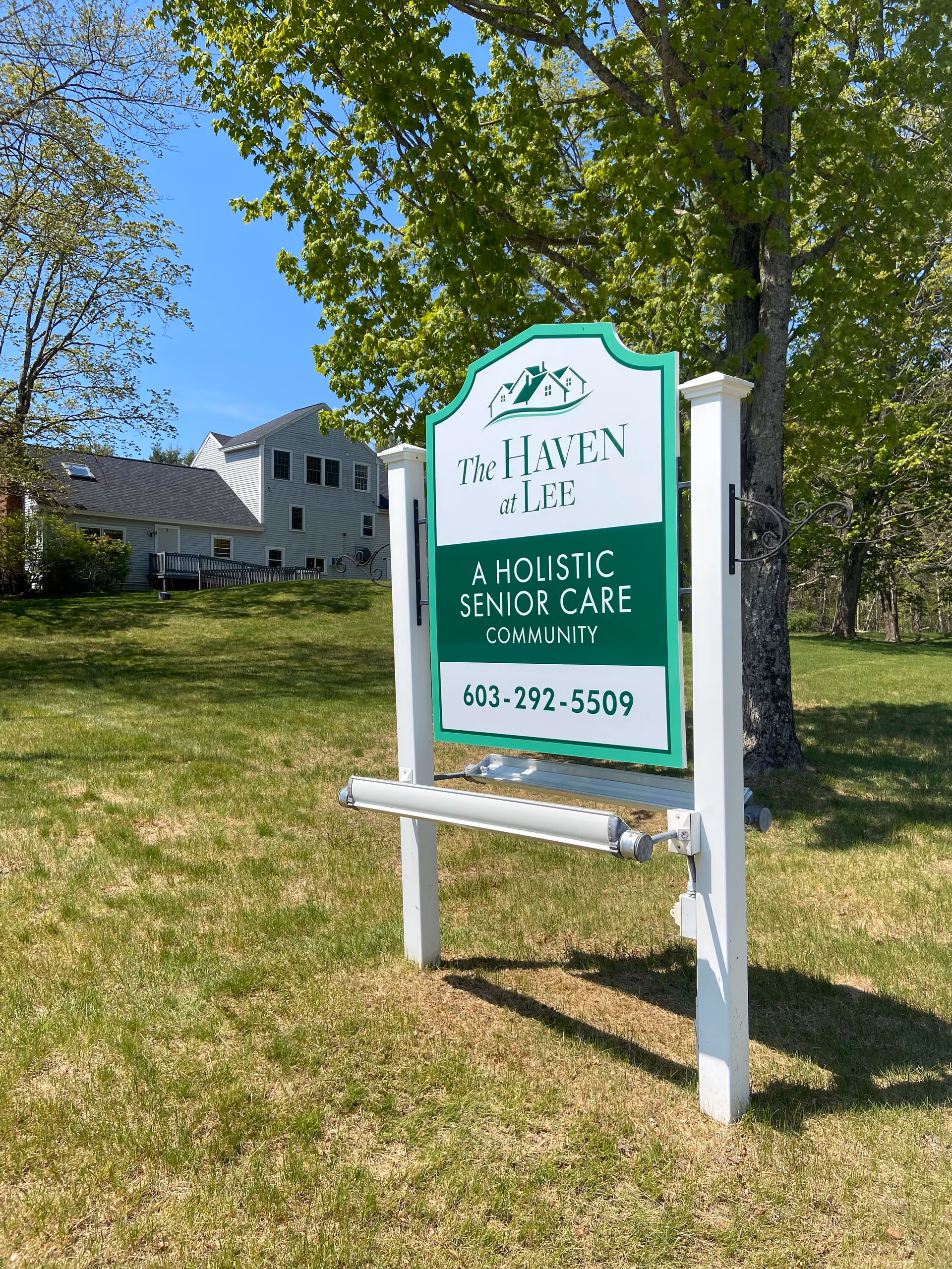 Outdoor view of a green and white sign for The Haven at Lee, a holistic senior care community, with a phone number 603-292-5509. The sign is set on a grassy area with trees and a building in the background under a clear blue sky.