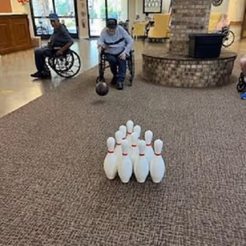 Two elderly individuals in wheelchairs playing indoor bowling in a common area with carpeted floor and a brick fireplace in the background.
