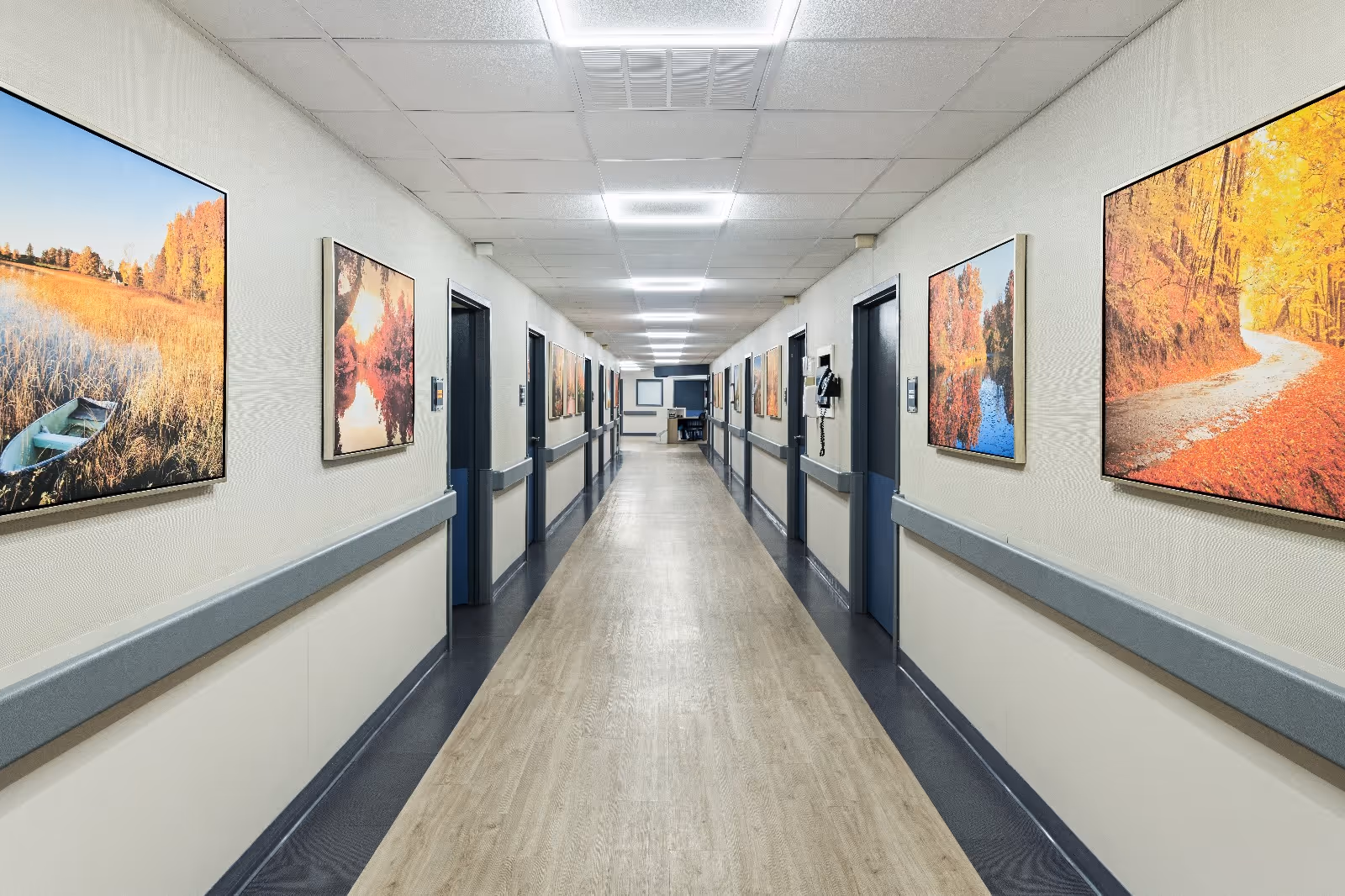 Long, well-lit interior hallway in a senior living facility lined with doors, handrails, and landscape paintings.