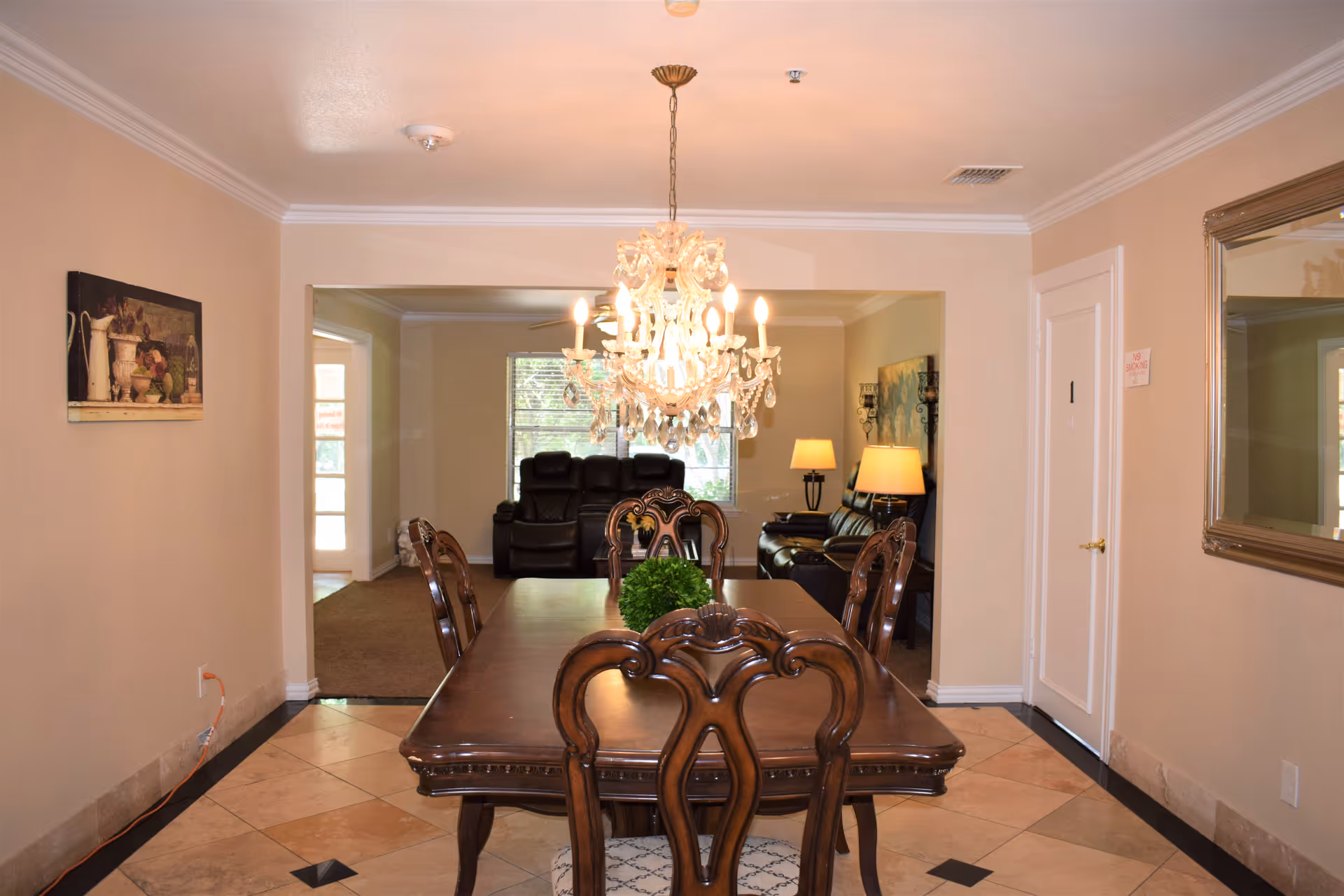 Interior view of a dining room with a wooden dining table and six chairs. A decorative chandelier hangs above the table. In the background, there is a living room area with leather sofas, a floor lamp, and a window letting in natural light. The walls are painted beige, and there is a large mirror on the right wall and a framed painting on the left wall.