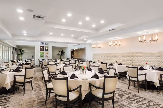 Bright, spacious dining room with multiple round tables set with white tablecloths, black napkins, and upholstered chairs under modern chandeliers.