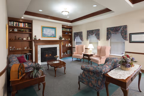 A cozy living room with floral upholstered sofas and two pink armchairs arranged around a wooden coffee table in front of a fireplace. Built-in wooden shelves flank the fireplace, holding books and decorative items. Three windows with patterned valances let in natural light, and a side table with a lamp is placed between the armchairs. A wooden console table with a lace runner and a floral arrangement is positioned near the foreground.