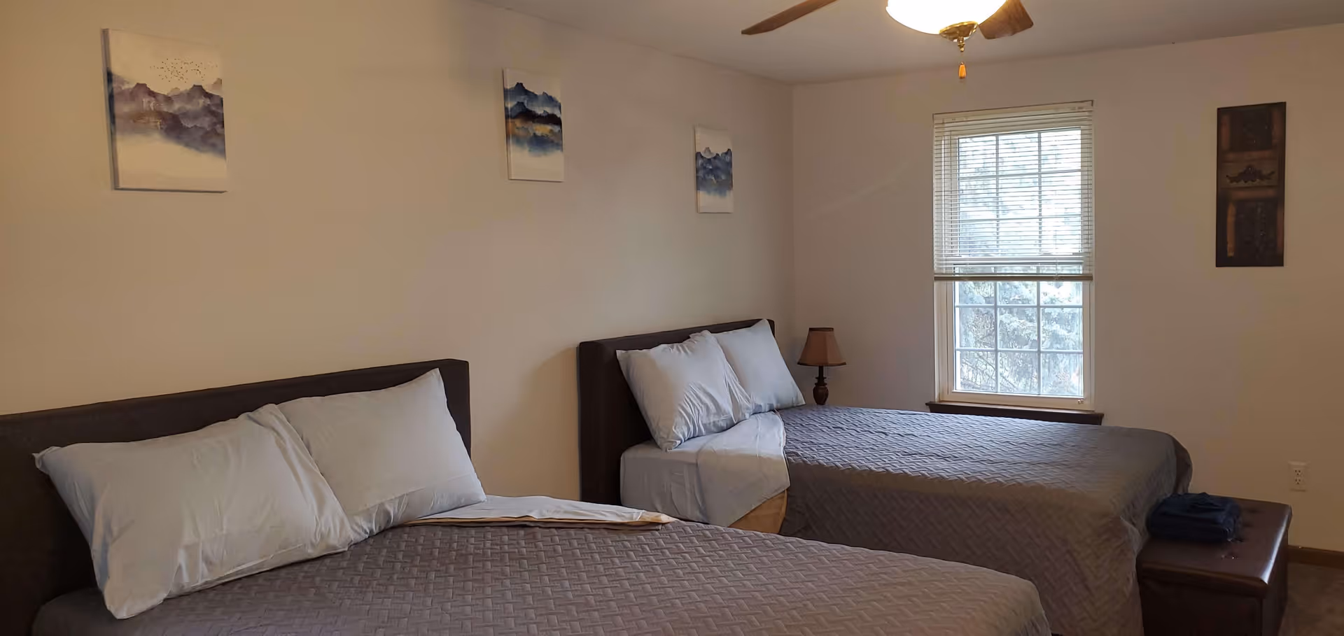 A bedroom with two beds covered in gray quilted bedspreads and white pillows. There is a window with blinds on the right wall, a small lamp on a nightstand between the beds, and four pieces of wall art above and beside the beds. A ceiling fan is partially visible at the top.
