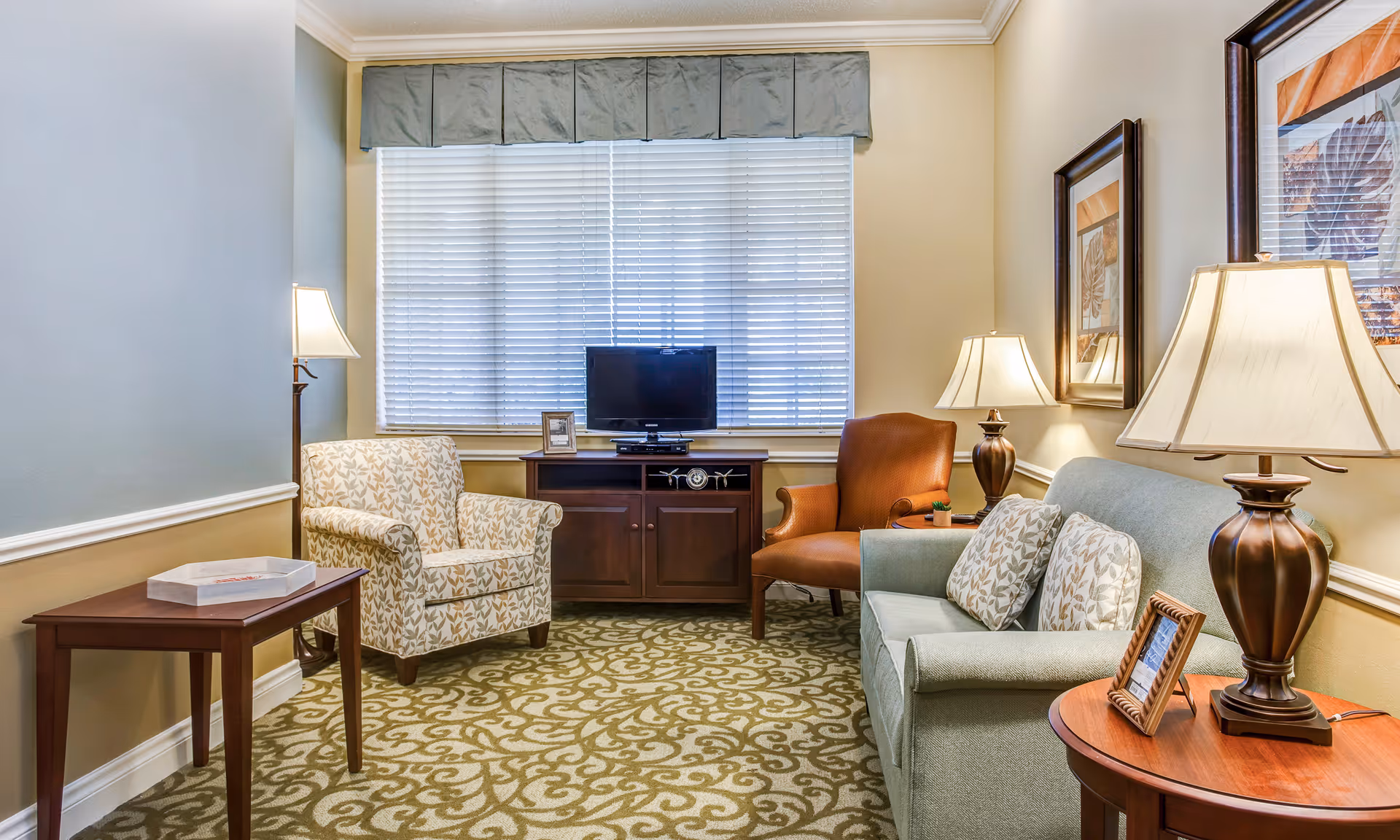 Bright, well-appointed living room with a sofa, patterned armchairs, side tables and lamps, a TV on a cabinet under a large window, and framed artwork.