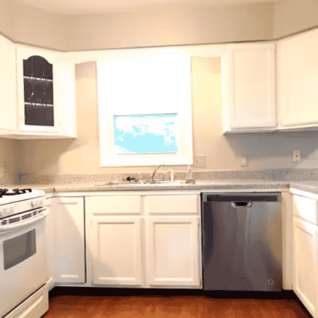A clean kitchen with white cabinets, a stainless steel dishwasher, a white gas stove, a countertop with a sink under a window, and beige walls.