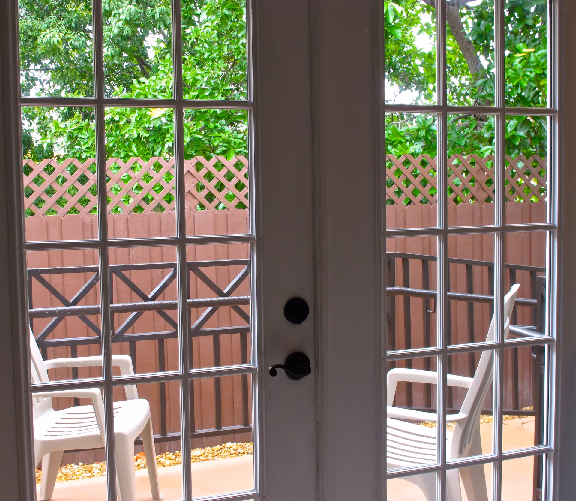 View through French doors onto a small patio with two white plastic chairs, a brown lattice fence and green foliage beyond.
