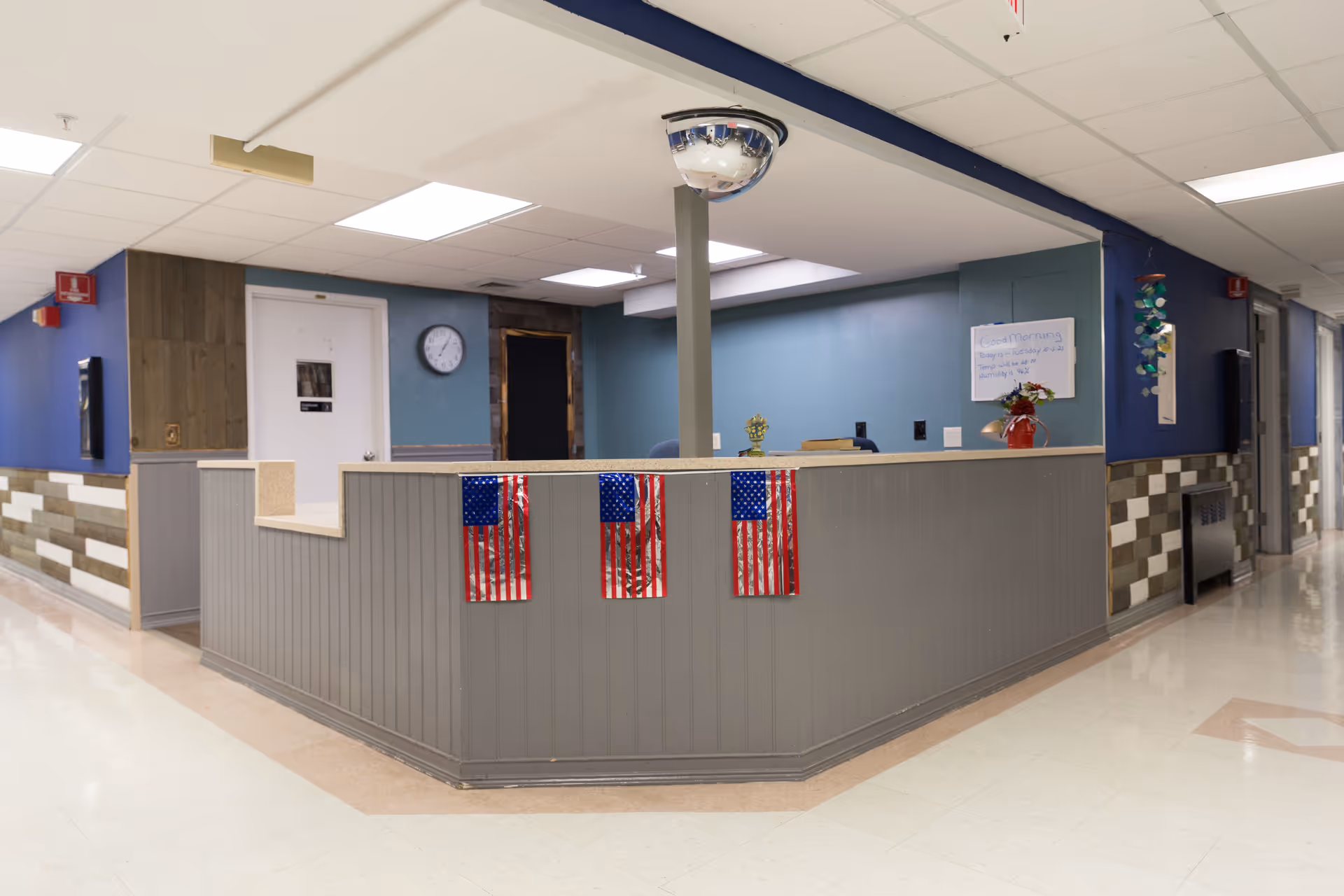 Reception desk area in a senior community facility with three small American flags attached to the front. The walls are painted blue and gray with a clock on the wall and a whiteboard displaying a morning message. There is a convex security mirror mounted on the ceiling above the desk. The hallway floors are tiled and the area is well-lit with ceiling lights.