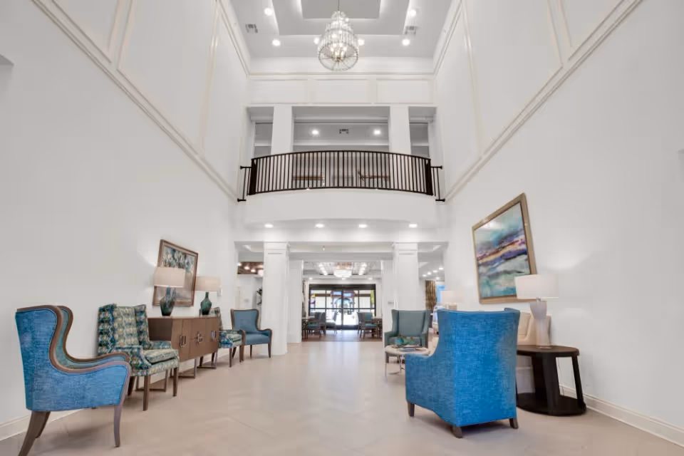 A spacious and bright lobby area with high ceilings and a chandelier. The room features blue and patterned armchairs arranged along the walls, wooden side tables with lamps, and framed artwork. A balcony with a railing overlooks the lobby, and large windows and doors are visible at the far end.