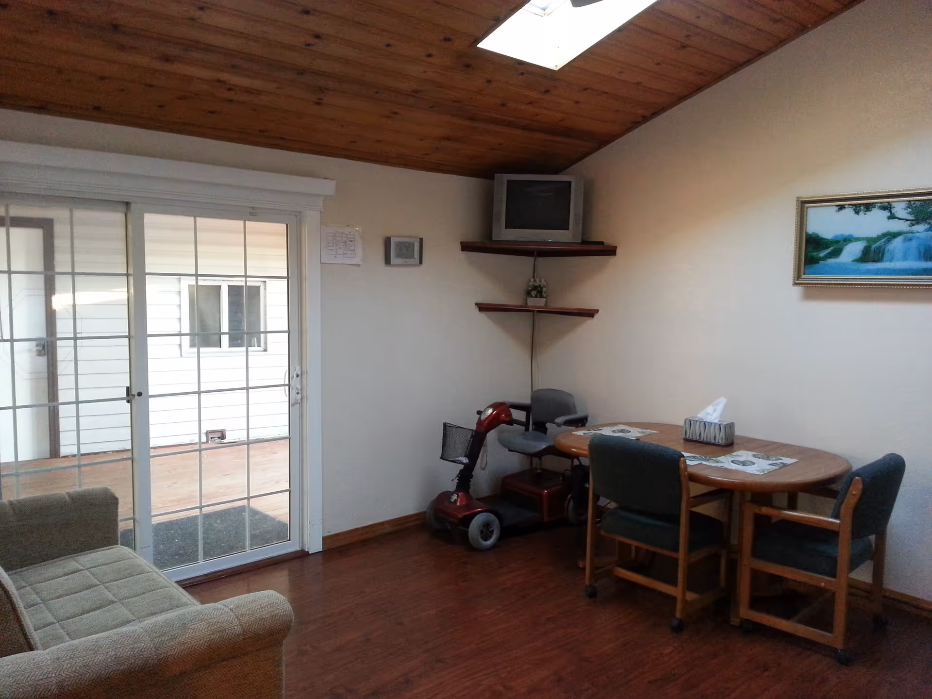 Room with sliding glass doors, a couch, round table with chairs, a mobility scooter, and a TV on corner shelves.